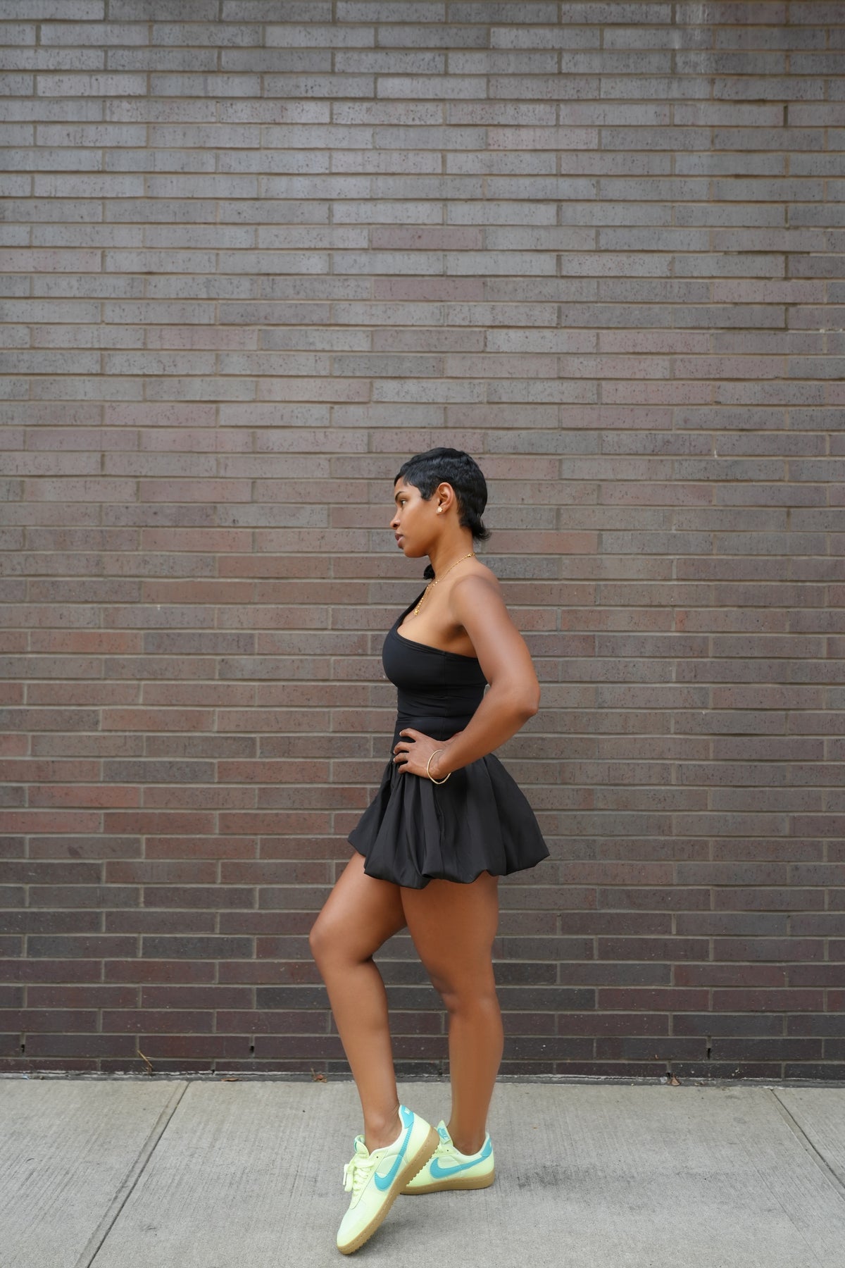 Woman in a black outfit with yellow shoes standing against a brick wall.